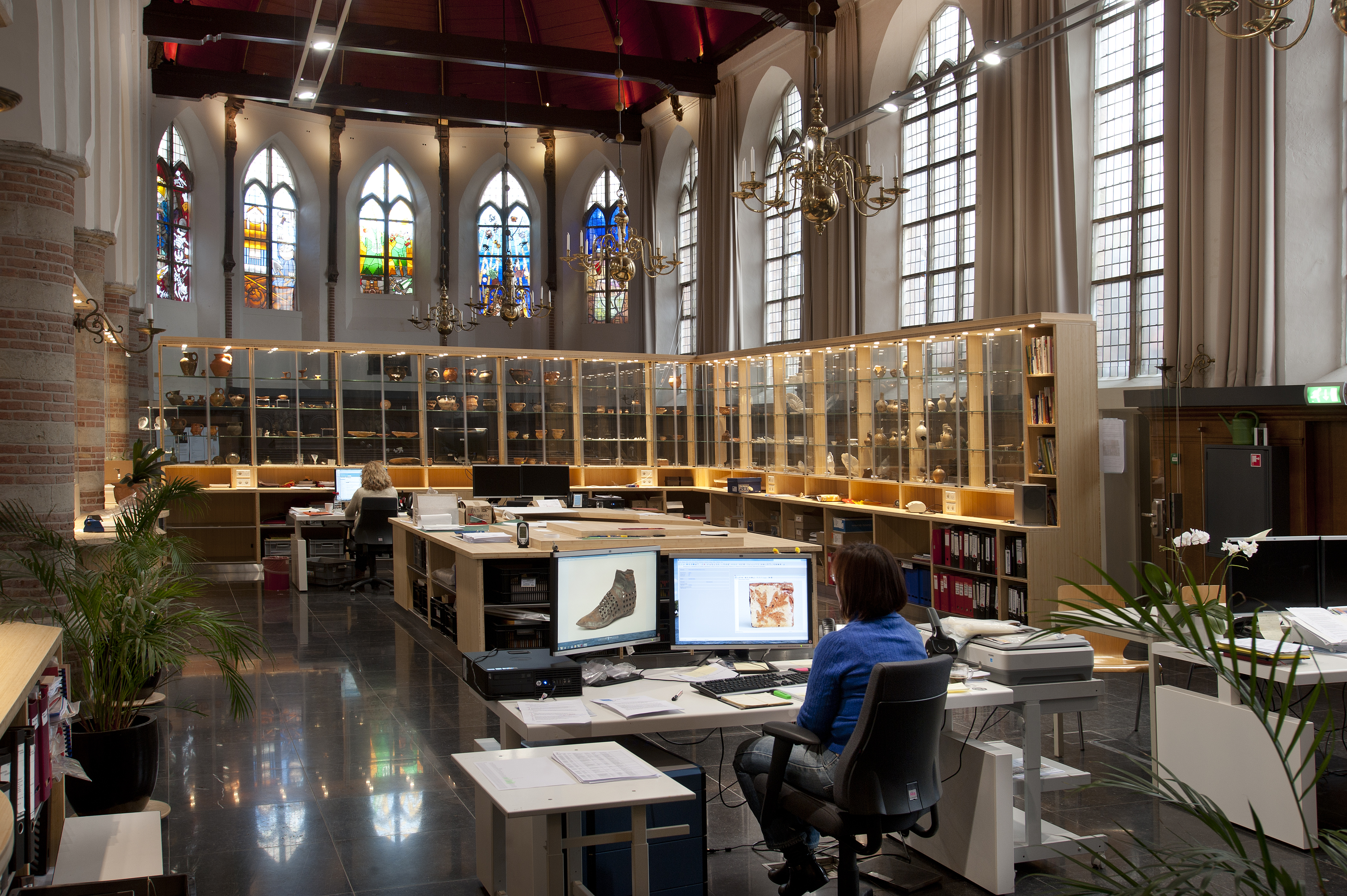 Op de achtergrond de glas-in-loodramen ontworpen door striptekenaars. De ramen laten het nieuwe gebruik van de kerk zien. De binnenruimte wordt namelijk gebruikt als werkruimte en tentoonstellingsruimte voor de Dienst Archeologie van Haarlem. 